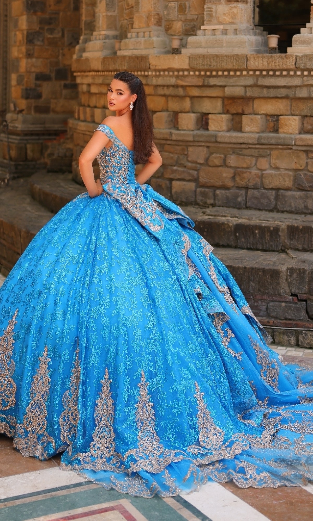 A woman poses by a stone wall wearing the Quinceanera Dress 54324 By Amarra—an elaborate blue, off-the-shoulder ball gown featuring gold and lace embroidery with a sweeping long train.
